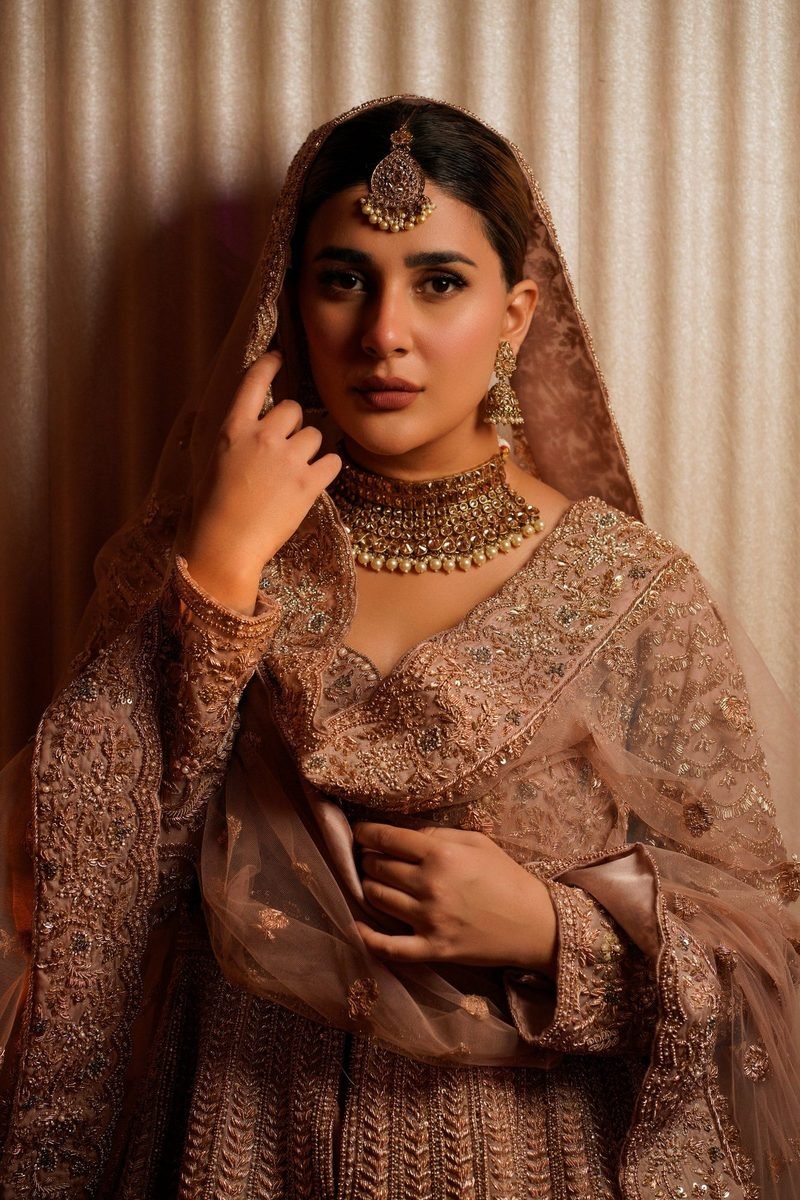 Bride in an embroidered outfit and sheer dupatta wearing a gold choker necklace, maang tikka and drop earrings, one hand raised near her face