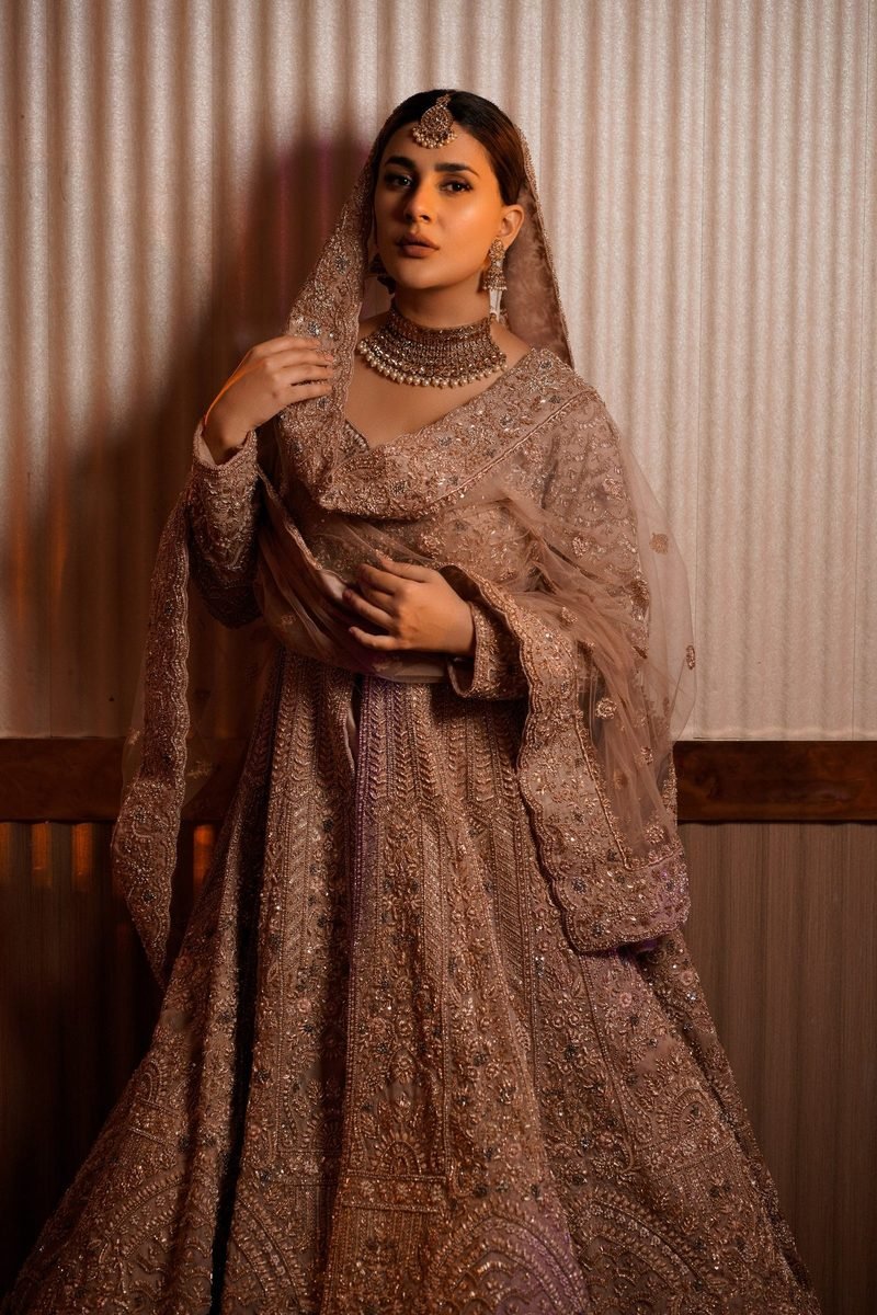 Full-length portrait of a bride in an embroidered lehenga and sheer dupatta, wearing a gold choker necklace and drop earrings, against a ribbed cream wall