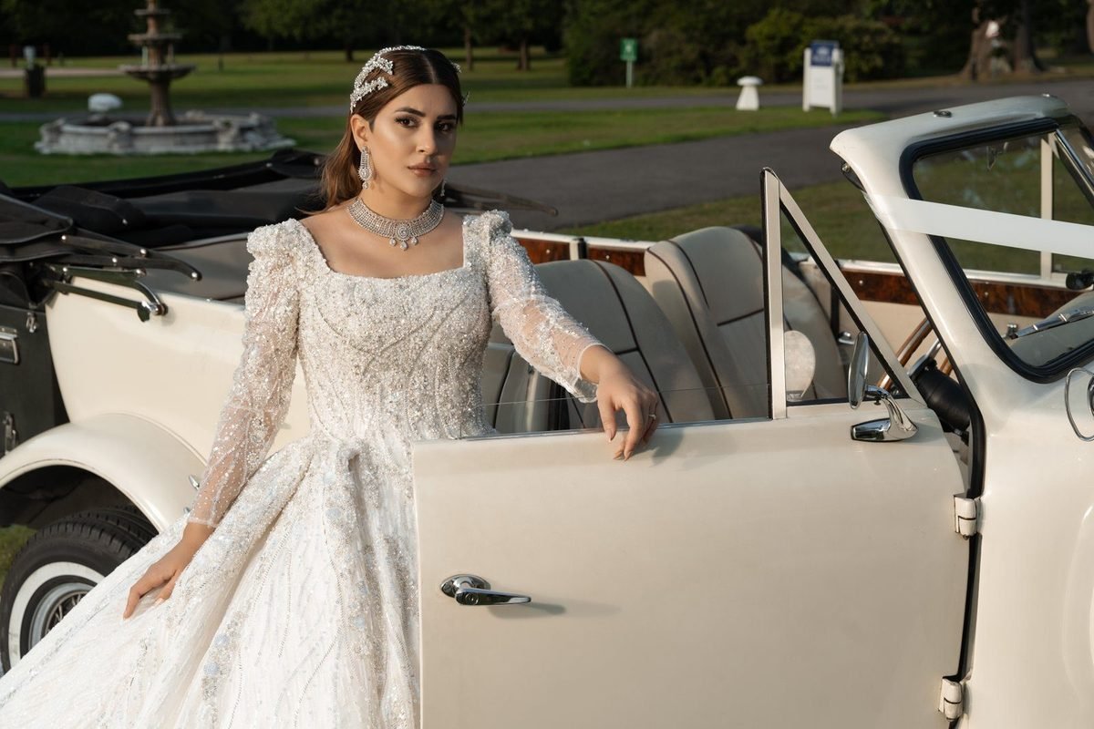 Bride in a white gown leaning on the door of a cream vintage car in a green garden with a fountain behind