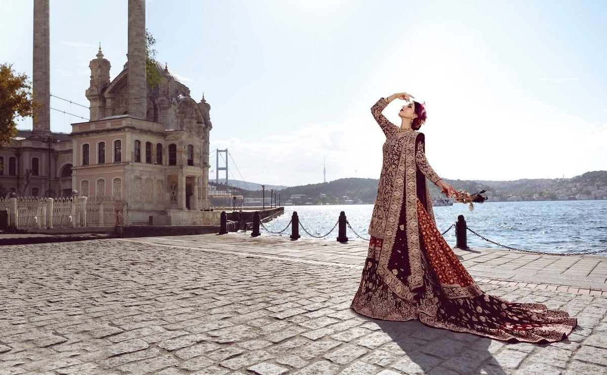 Bride in a red and gold lehenga posing on a sunlit waterfront promenade under a bright sky