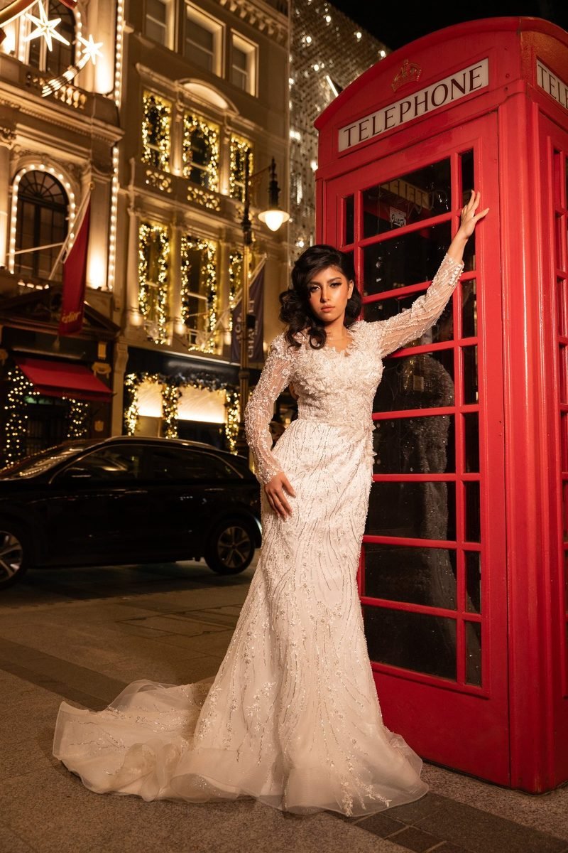 Bride in a fitted white gown posing beside a red telephone box on a lit street at dusk