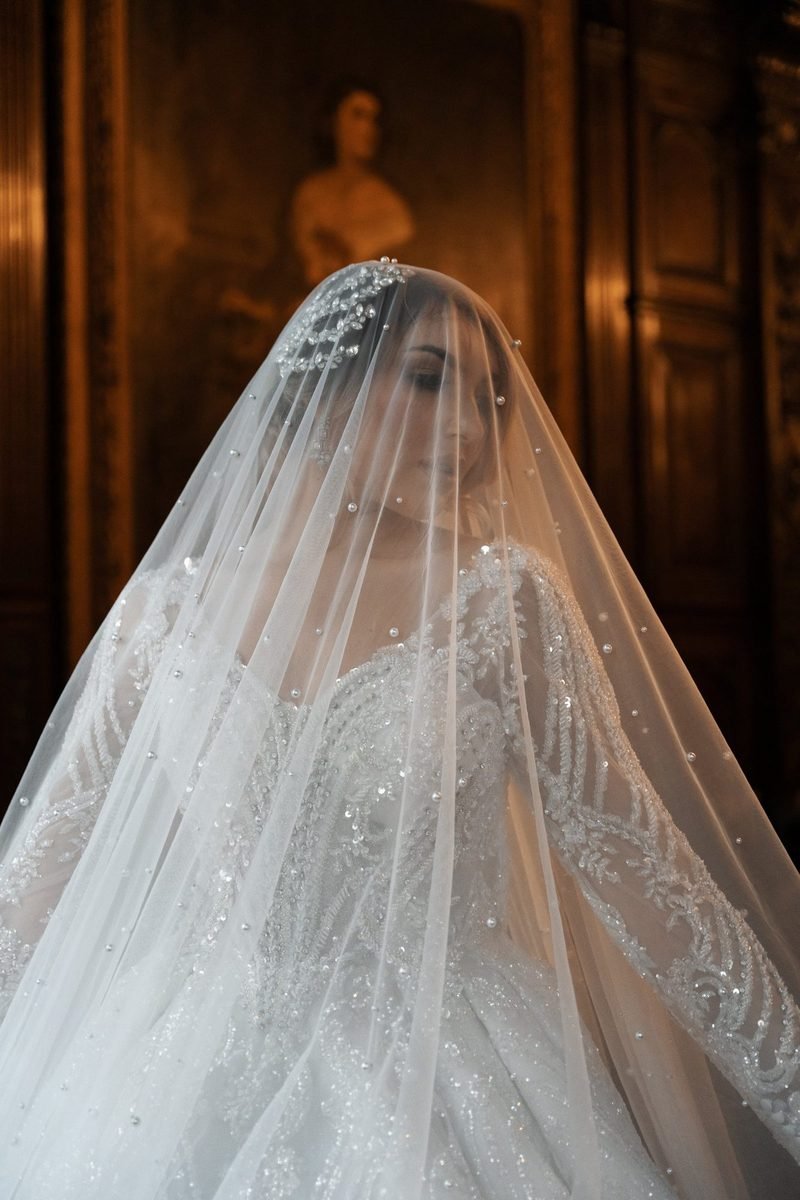 Bride veiled in sheer tulle and a beaded gown, softly lit against a dark period interior