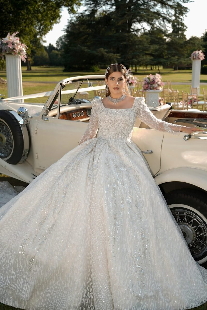 Bride in a white beaded ballgown standing beside a ribbon-decked cream vintage car, with flower stands and chairs on a sunlit lawn behind