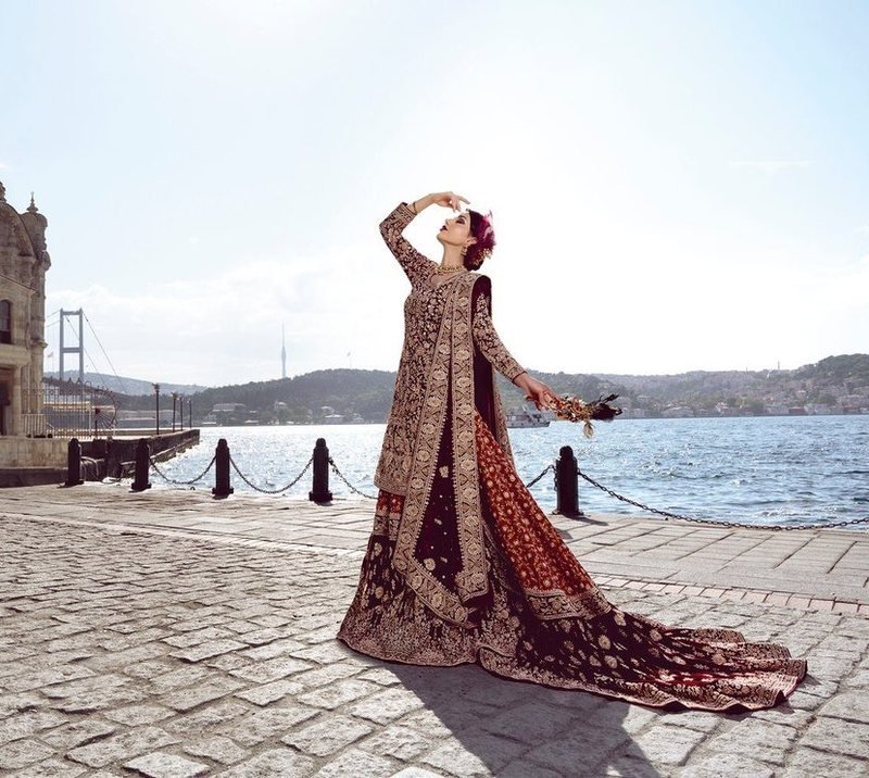 Bride in a red and gold lehenga posing on a sunlit waterfront promenade under a bright sky