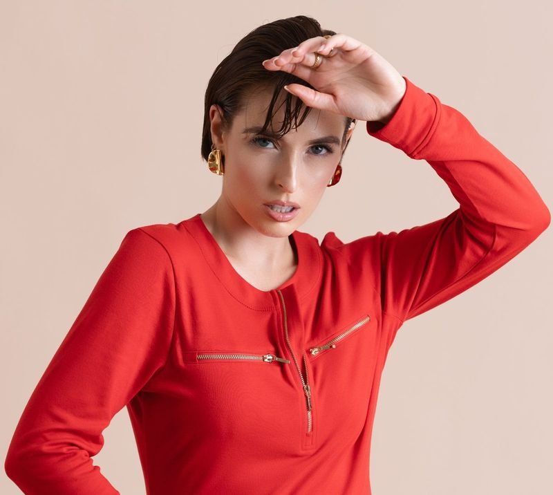 Woman in a fitted red dress with one hand raised to her forehead, against a soft pink backdrop