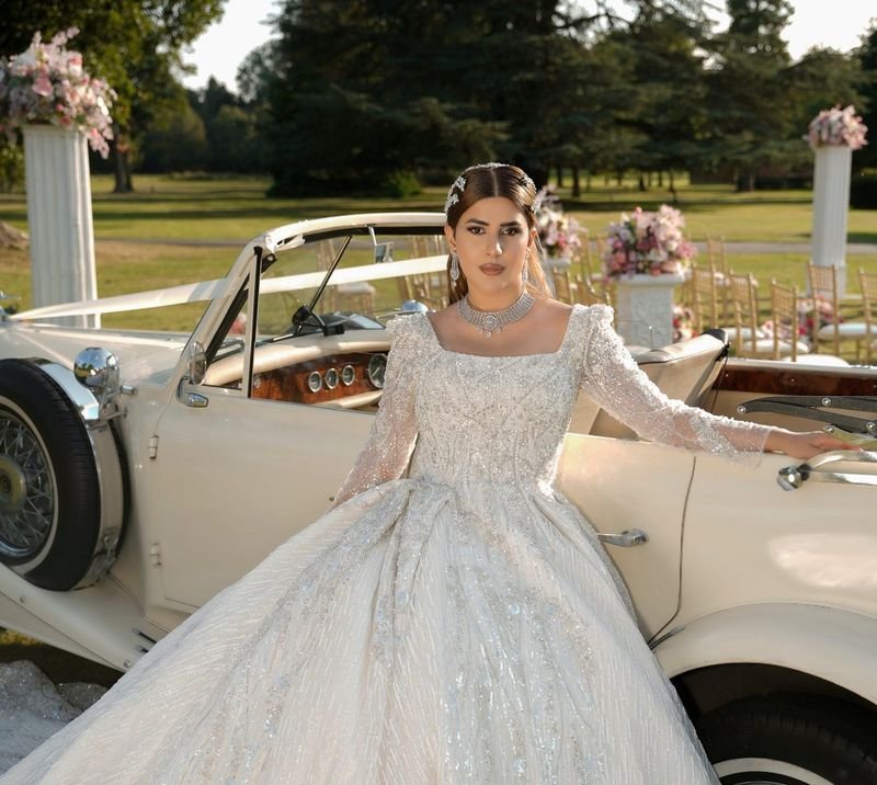 Bride in a white beaded ballgown standing beside a ribbon-decked cream vintage car on a sunlit lawn