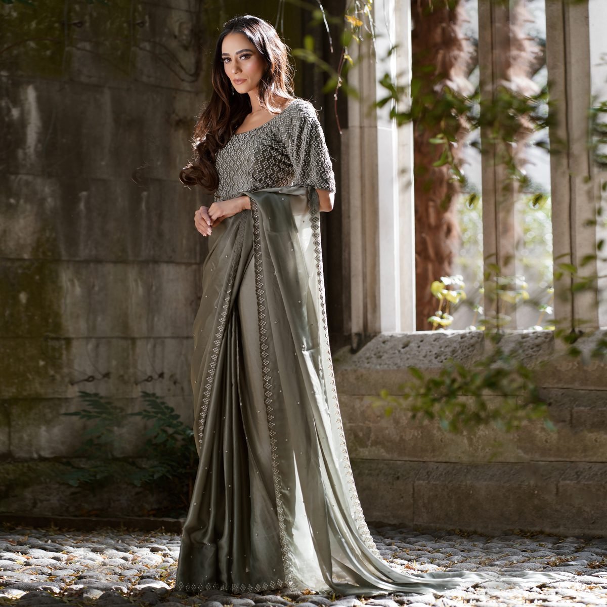 Model in a grey-green saree standing at a stone window frame with sunlit greenery behind