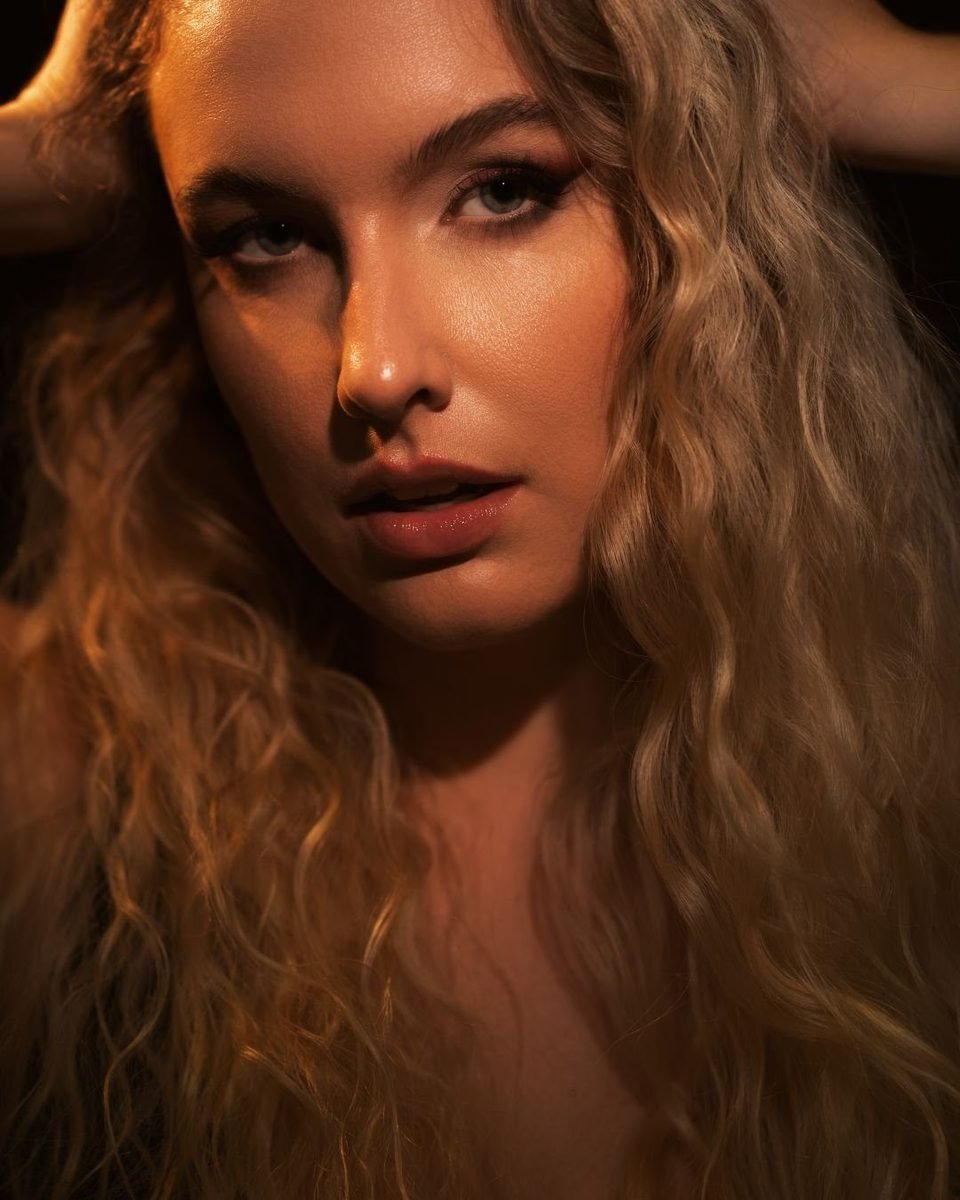 Close portrait of a woman with long curly hair in warm, low light, looking into the lens