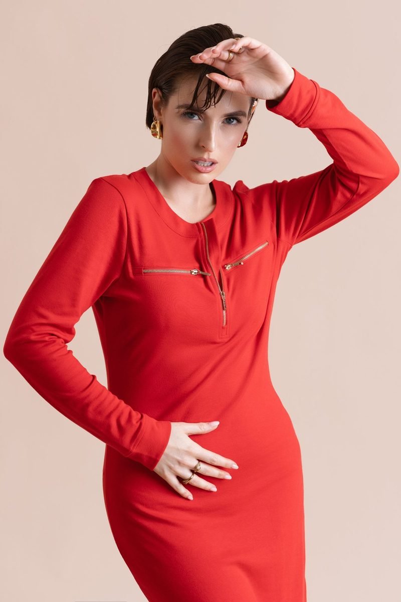 Woman in a fitted red dress with one hand raised to her forehead and the other on her hip, against a soft pink backdrop