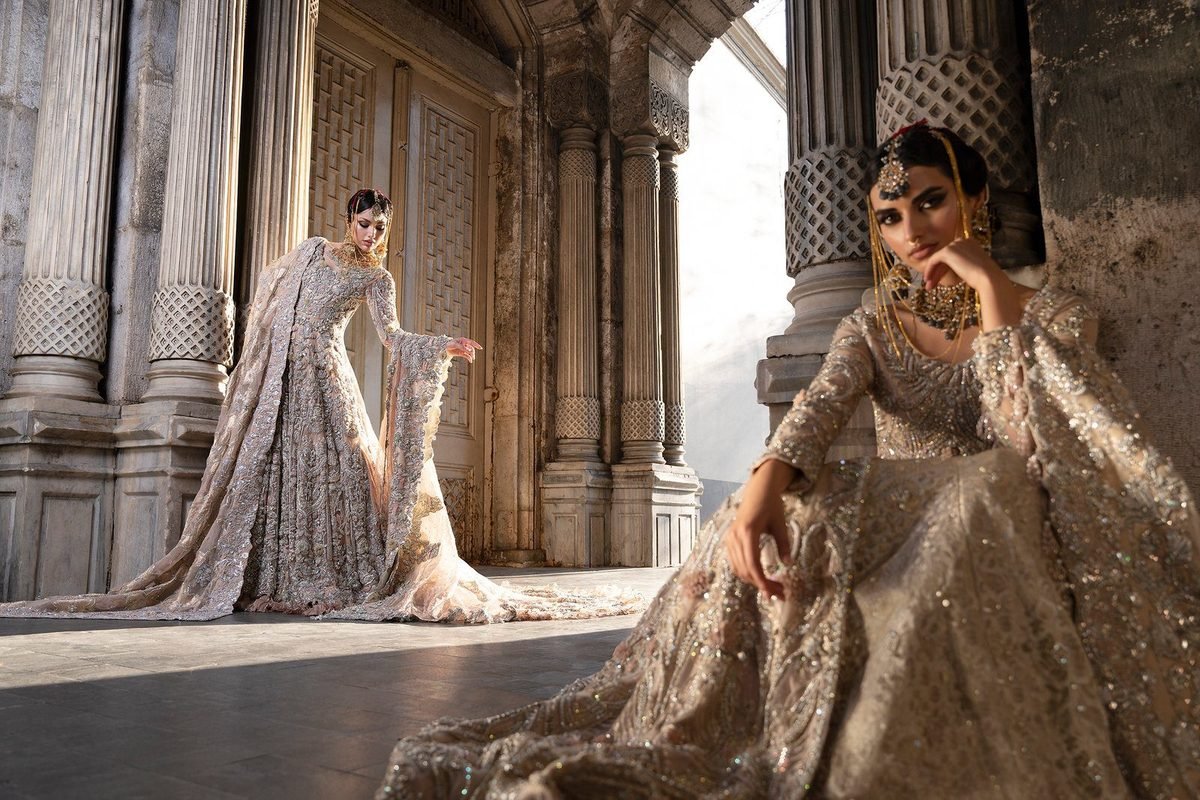 Two models in embroidered gowns posed between tall stone columns in an arched hall