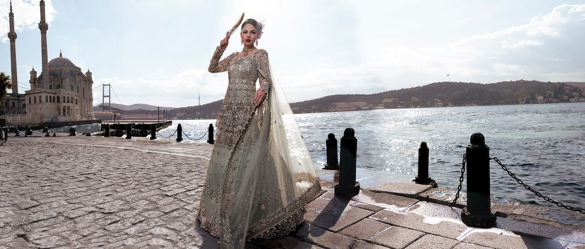 Bride in a pale embroidered lehenga standing on a waterfront promenade with a mosque in the background