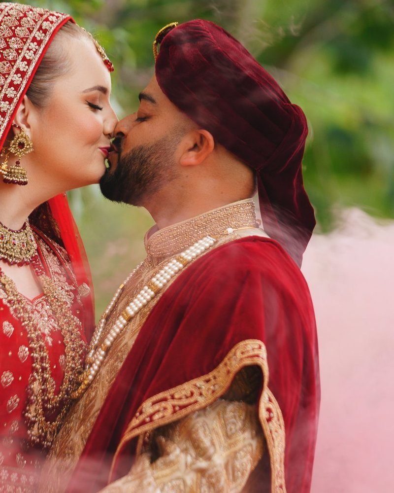 Bride and groom in red and gold Asian wedding outfits sharing a kiss with pink smoke behind them