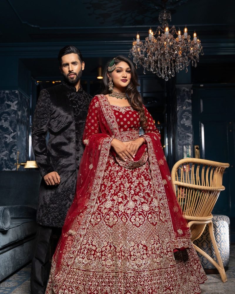 Couple in a red embroidered lehenga and a black velvet sherwani beneath a chandelier in a teal room