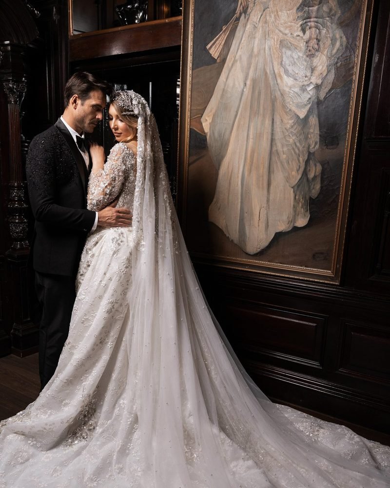 Bride in a long veil and groom in a black tuxedo embracing in front of a large framed painting