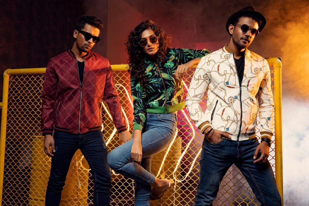 Three models in bomber jackets and sunglasses in front of a fence with smoke and orange light