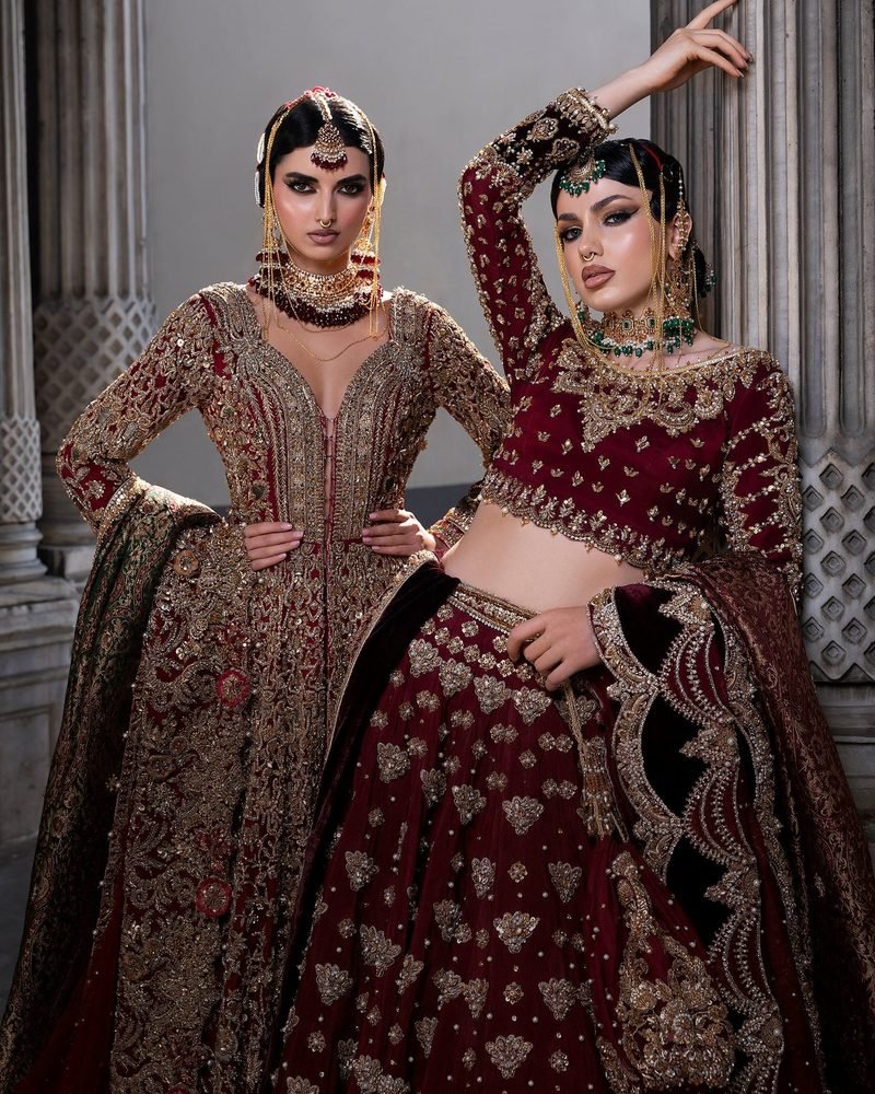 Two brides in maroon and gold embroidered lehengas posing between stone columns