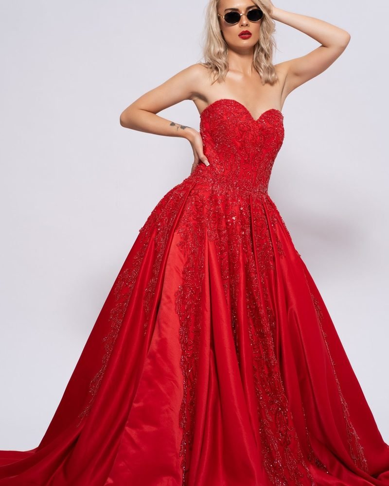 Model in a strapless red ball gown with a long train and one hand raised to her head